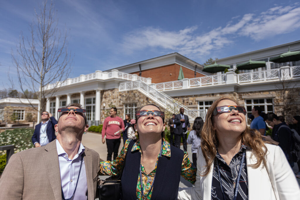 One man and two women in suits look at the sky with eclipse glasses.