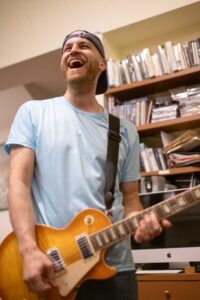 Photo of a man laughing and playing guitar in front of a bookshelf.