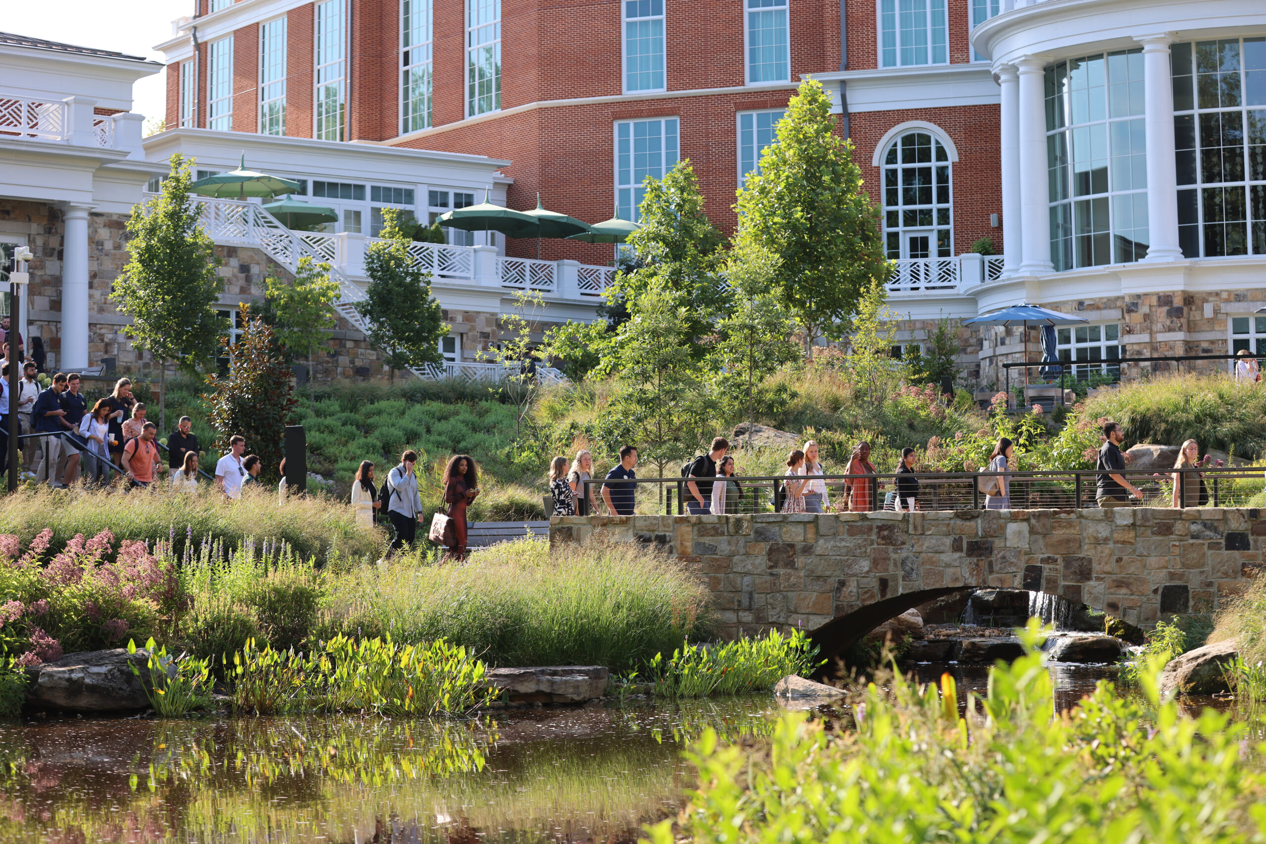 Students walking across a bridge in a garden with The Forum Hotel behind them.