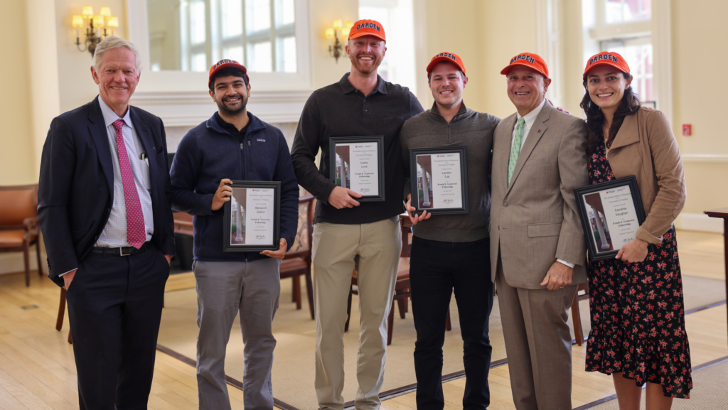 Frank Genovese and Dean Scott Beardsley with the 2025 Genovese Fellows, each holding framed certificates and wearing bright orange Darden hats.
